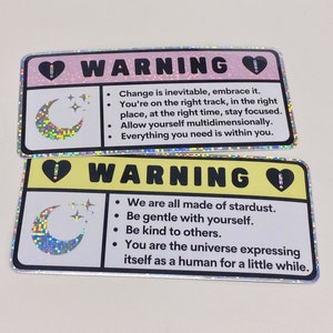 Puede incluir: Dos pegatinas hologr&aacute;ficas con un dise&ntilde;o de luna creciente y estrellas. Las pegatinas tienen un fondo amarillo y rosa con texto negro que dice "ADVERTENCIA". El texto en la pegatina rosa dice "El cambio es inevitable, ac&eacute;ptalo. Est&aacute;s en el camino correcto, en el lugar correcto, en el momento correcto, mantente concentrado. Perm&iacute;tete ser multidimensional. Todo lo que necesitas est&aacute; dentro de ti." El texto en la pegatina amarilla dice "Todos estamos hechos de polvo de estrellas. S&eacute; amable contigo mismo. S&eacute; amable con los dem&aacute;s. T&uacute; eres el universo expres&aacute;ndose como un humano por un rato."