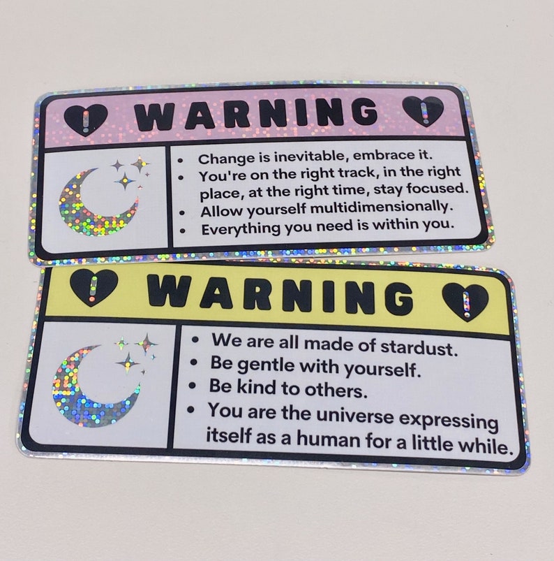 Puede incluir: Dos pegatinas hologr&aacute;ficas con un dise&ntilde;o de luna creciente y estrellas. Las pegatinas tienen un fondo amarillo y rosa con texto negro que dice "ADVERTENCIA". El texto en la pegatina rosa dice "El cambio es inevitable, ac&eacute;ptalo. Est&aacute;s en el camino correcto, en el lugar correcto, en el momento correcto, mantente concentrado. Perm&iacute;tete ser multidimensional. Todo lo que necesitas est&aacute; dentro de ti." El texto en la pegatina amarilla dice "Todos estamos hechos de polvo de estrellas. S&eacute; amable contigo mismo. S&eacute; amable con los dem&aacute;s. T&uacute; eres el universo expres&aacute;ndose como un humano por un rato."