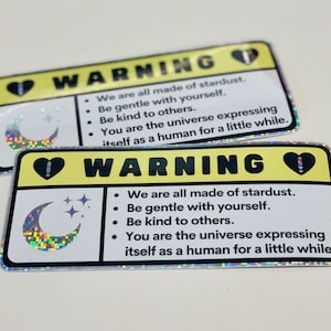 Puede incluir: Dos pegatinas de advertencia amarillas con un coraz&oacute;n negro y un signo de exclamaci&oacute;n, una luna creciente con estrellas y el texto "WARNING" y "We are all made of stardust. Be gentle with yourself. Be kind to others. You are the universe expressing itself as a human for a little while."
