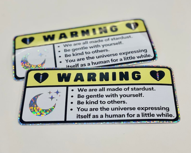 Puede incluir: Dos pegatinas de advertencia amarillas con un coraz&oacute;n negro y un signo de exclamaci&oacute;n, una luna creciente con estrellas y el texto "WARNING" y "We are all made of stardust. Be gentle with yourself. Be kind to others. You are the universe expressing itself as a human for a little while."