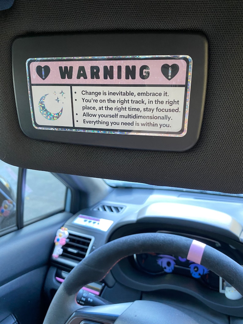 Puede incluir: Una visera de coche rosa y blanca con un cartel de advertencia que dice "WARNING" con un coraz&oacute;n a cada lado. El cartel tambi&eacute;n incluye una luna creciente con estrellas y el texto "Change is inevitable, embrace it. You're on the right track, in the right place, at the right time, stay focused. Allow yourself multidimensionally. Everything you need is within you."