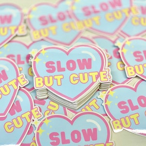 May include: A stack of heart-shaped stickers with the text "Slow But Cute" in pink and yellow letters on a light blue background. The stickers have a sparkly finish.