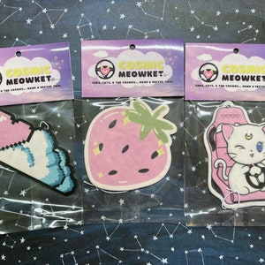 May include: Three car air fresheners with a pink and blue color scheme. The designs are a pixelated heart and cloud, a strawberry, and a white cat in a car seat. The text on the air fresheners reads "Cosmic Meowket Cars, Cats, & The Cosmos... Make a Better Trip."