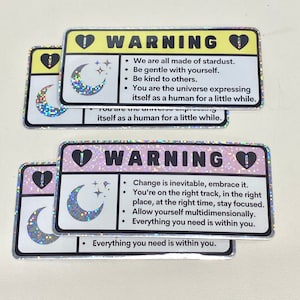 Puede incluir: Dos pegatinas hologr&aacute;ficas con un dise&ntilde;o de luna creciente y estrellas. Las pegatinas tienen un fondo rosa y amarillo con texto negro que dice "ADVERTENCIA" e incluye mensajes inspiradores. El texto en la pegatina amarilla dice: "Todos estamos hechos de polvo de estrellas. S&eacute; amable contigo mismo. S&eacute; amable con los dem&aacute;s. T&uacute; eres el universo que se expresa como un humano por un tiempo." El texto en la pegatina rosa dice: "El cambio es inevitable, ac&eacute;ptalo. Est&aacute;s en el camino correcto, en el lugar correcto, en el momento correcto, mantente enfocado. Perm&iacute;tete ser multidimensional. Todo lo que necesitas est&aacute; dentro de ti."