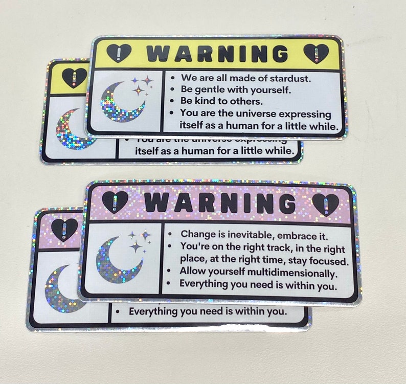 Puede incluir: Dos pegatinas hologr&aacute;ficas con un dise&ntilde;o de luna creciente y estrellas. Las pegatinas tienen un fondo rosa y amarillo con texto negro que dice "ADVERTENCIA" e incluye mensajes inspiradores. El texto en la pegatina amarilla dice: "Todos estamos hechos de polvo de estrellas. S&eacute; amable contigo mismo. S&eacute; amable con los dem&aacute;s. T&uacute; eres el universo que se expresa como un humano por un tiempo." El texto en la pegatina rosa dice: "El cambio es inevitable, ac&eacute;ptalo. Est&aacute;s en el camino correcto, en el lugar correcto, en el momento correcto, mantente enfocado. Perm&iacute;tete ser multidimensional. Todo lo que necesitas est&aacute; dentro de ti."