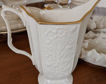 May include: A white porcelain pitcher with a gold rim and floral design. The pitcher has a handle and a wide base.