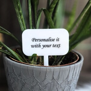 Peut inclure: Un marqueur de plante blanc aux coins arrondis, affichant le texte "Personalise it with your text" dans une écriture élégante. Le marqueur est placé dans un pot gris texturé avec une plante verte. La plante a de longues feuilles vertes.