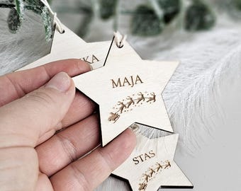 May include: Three light-colored wooden star-shaped ornaments featuring laser-engraved text and graphics. Each star displays a name, such as MAJA or STAS, above an engraved illustration of a sleigh pulled by reindeer. Each ornament includes a white string.