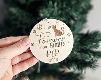May include: A round, light-colored wooden ornament featuring laser-engraved details including a cat silhouette, paw prints, and snowflakes. The text reads Forever in our HEARTS, PIP, and the year 2019. A small hole at the top allows for hanging.