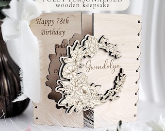 May include: A light-colored wooden keepsake card featuring a laser-cut floral wreath design and the name Gwendolyn. The card is engraved with the text Happy 78th Birthday and is tied with a white fabric ribbon on the left side with visible stitching.