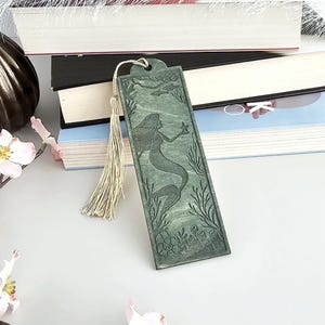 Könnte beinhalten: Ein grüner Holz-Lesezeichen mit einem Meerjungfrau-Design, das einen Seestern hält, mit einem Quastenanhänger. Das Lesezeichen ist rechteckig mit einem Loch oben. Die Meerjungfrau ist von Wasserpflanzen und Fischen umgeben.