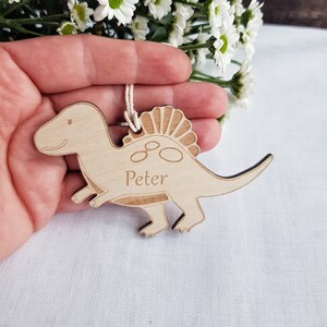 Könnte beinhalten: Holz-Dinosaurier-Anhänger mit einem eingravierten Namen. Der Dinosaurier ist braun und hat einen stacheligen Rücken. Der Name "Peter" ist in den Anhänger eingraviert.