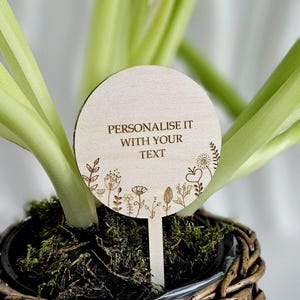 Peut inclure: Un marqueur de plante rond en bois clair avec le texte "PERSONNALISEZ-LE AVEC VOTRE TEXTE" gravé. Le marqueur présente un motif floral et est placé dans une plante en pot avec des feuilles vertes.