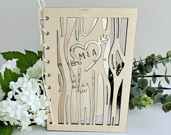 May include: A wooden wedding card featuring a heart design with the initials "M&A". The card has a tree-like cutout pattern and is bound with a white string. The background includes white flowers and green foliage.