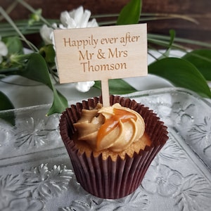May include: A cupcake with caramel frosting and a wooden sign that reads "Happily ever after Mr & Mrs Thomson". The cupcake is in a brown paper liner and sits on a clear glass plate. White flowers and green leaves are in the background.