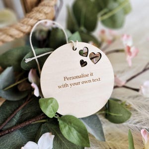May include: A circular light-coloured wooden ornament featuring three heart-shaped cutouts at the top and a white string loop for hanging. The centre of the wooden disc is engraved with the text, Personalise it with your own text, in a simple font.