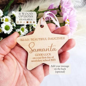 May include: A star-shaped light wood ornament with a pink ribbon loop. The text reads: TO MY BEAUTIFUL DAUGHTER Samanta GOOD LUCK on your first day of secondary school 2023. An overlay box states 6 WOOD OPTIONS AVAILABLE and another states Add your message on the back.
