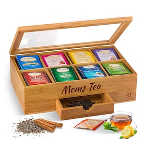 May include: A bamboo tea box with a glass lid, containing various Twinings tea bags. The box has a drawer with loose tea and the words "Moms Tea" engraved on the front. A cup of tea, lemon slices, and cinnamon sticks are also visible.