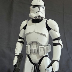 May include: A white and black action figure of a Clone Trooper from Star Wars. The figure is wearing a white helmet and armor, and is holding a black blaster rifle. The figure is standing on a black stand.