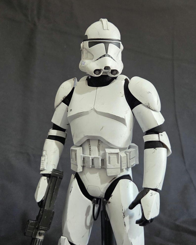 May include: A white and black action figure of a Clone Trooper from Star Wars. The figure is wearing a white helmet and armor, and is holding a black blaster rifle. The figure is standing on a black stand.
