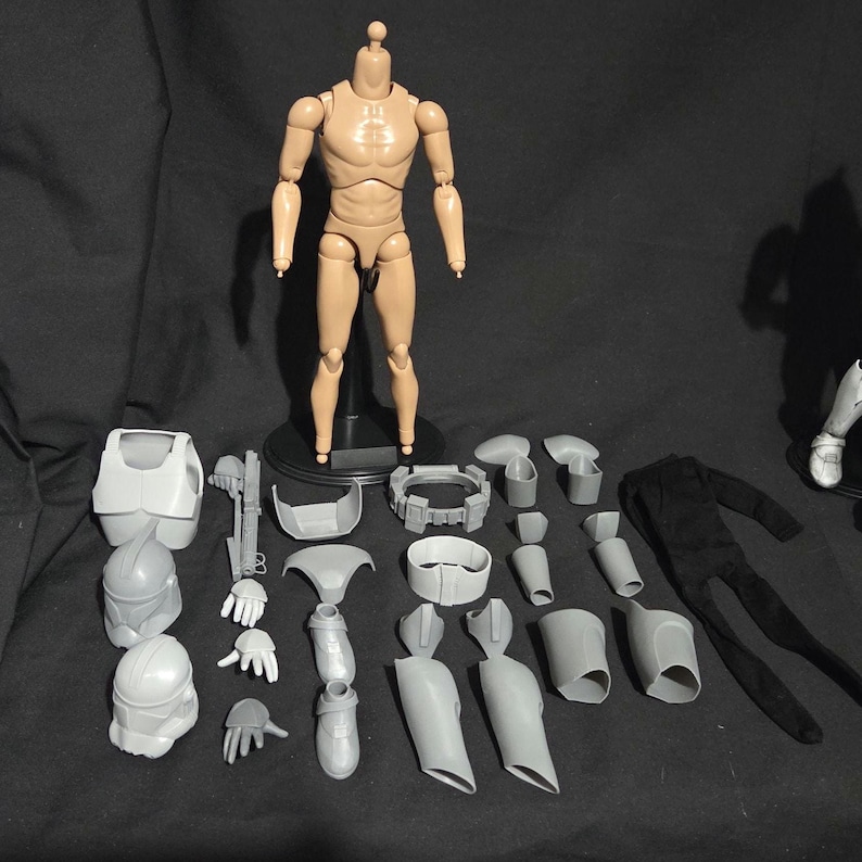 May include: A tan articulated action figure with various gray and white accessories, including helmets, armor, and clothing. The figure stands on a black base, with additional parts arranged around it.