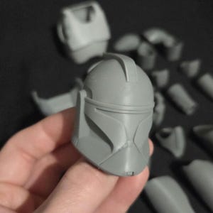 May include: A gray, 3D-printed helmet, held in a hand, with various detached parts scattered in the background. The helmet features a visor and distinct facial details, suggesting a sci-fi or cosplay theme.