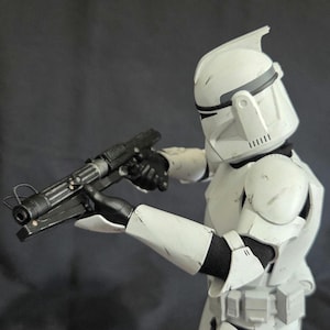 May include: A white and black action figure of a soldier in a helmet and armor, holding a black blaster rifle. The figure is in a combat stance, with the weapon pointed forward. The armor has some weathering.