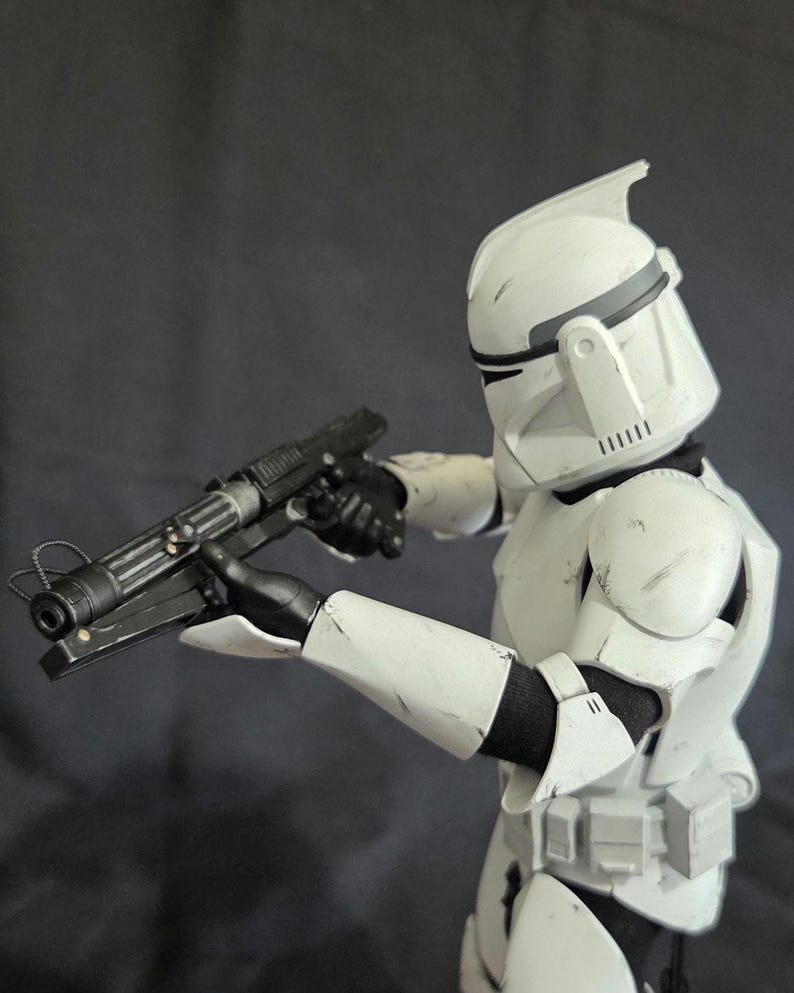 May include: A white and black action figure of a soldier in a helmet and armor, holding a black blaster rifle. The figure is in a combat stance, with the weapon pointed forward. The armor has some weathering.