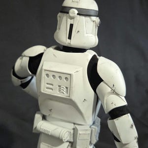 May include: A white action figure of a clone trooper from a science fiction franchise. The figure is wearing white armor with black accents and a helmet. The figure is posed with its back to the viewer.