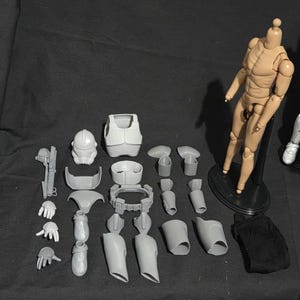 May include: A collection of unpainted, gray plastic action figure parts, including a helmet, armor, limbs, and accessories. A tan-colored articulated figure stands on a black base. A black fabric piece is also included.