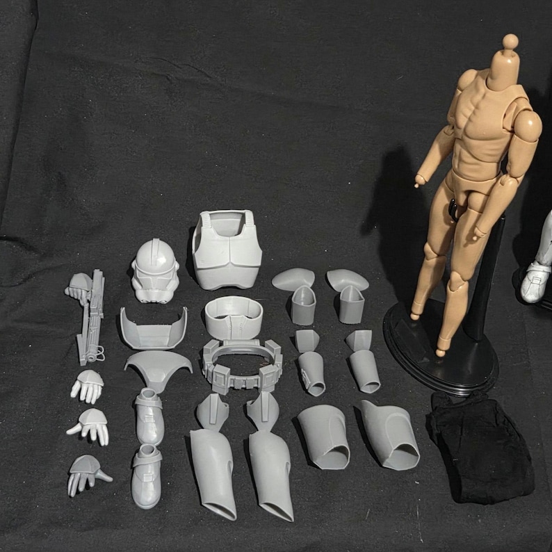 May include: A collection of unpainted, gray plastic action figure parts, including a helmet, armor, limbs, and accessories. A tan-colored articulated figure stands on a black base. A black fabric piece is also included.