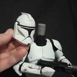 May include: A white and black action figure of a clone trooper from Star Wars. The figure is holding the helmet in one hand. The figure has a black weapon. The armor is white with black accents and some weathering.