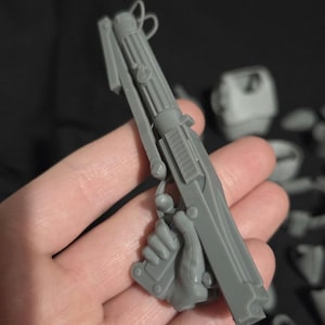 May include: A gray plastic model of a blaster pistol, held in a hand. The pistol has a long barrel with ribbed detailing, a trigger, and a handle. The model is part of a larger set of components.