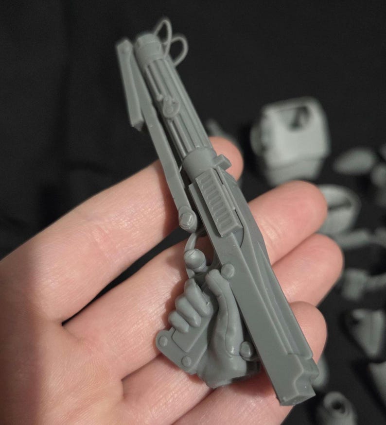 May include: A gray plastic model of a blaster pistol, held in a hand. The pistol has a long barrel with ribbed detailing, a trigger, and a handle. The model is part of a larger set of components.