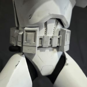 May include: Close-up of a white and gray stormtrooper armor torso. The armor features a detailed belt with rectangular panels and a chest piece with a distinct design. The surface shows signs of wear, suggesting a battle-worn appearance.
