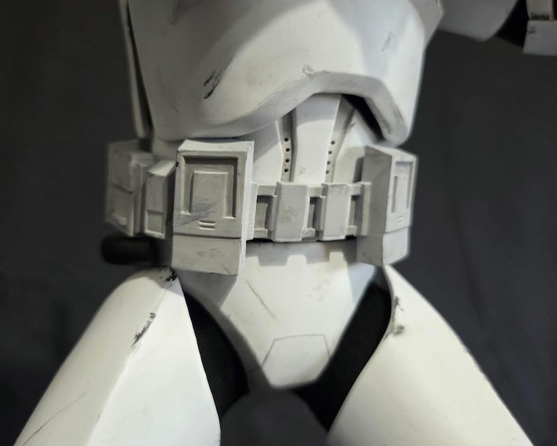May include: Close-up of a white and gray stormtrooper armor torso. The armor features a detailed belt with rectangular panels and a chest piece with a distinct design. The surface shows signs of wear, suggesting a battle-worn appearance.