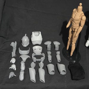 May include: A disassembled action figure with various gray plastic armor pieces, a helmet, and a tan articulated body. The figure is posed on a black stand, with additional accessories and a black fabric piece.
