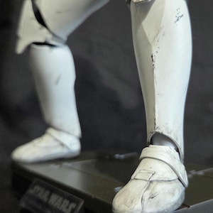 May include: Close-up of a white Stormtrooper action figure's legs and feet. The figure is detailed with battle-worn markings and is posed on a black base. The base has the Star Wars logo.