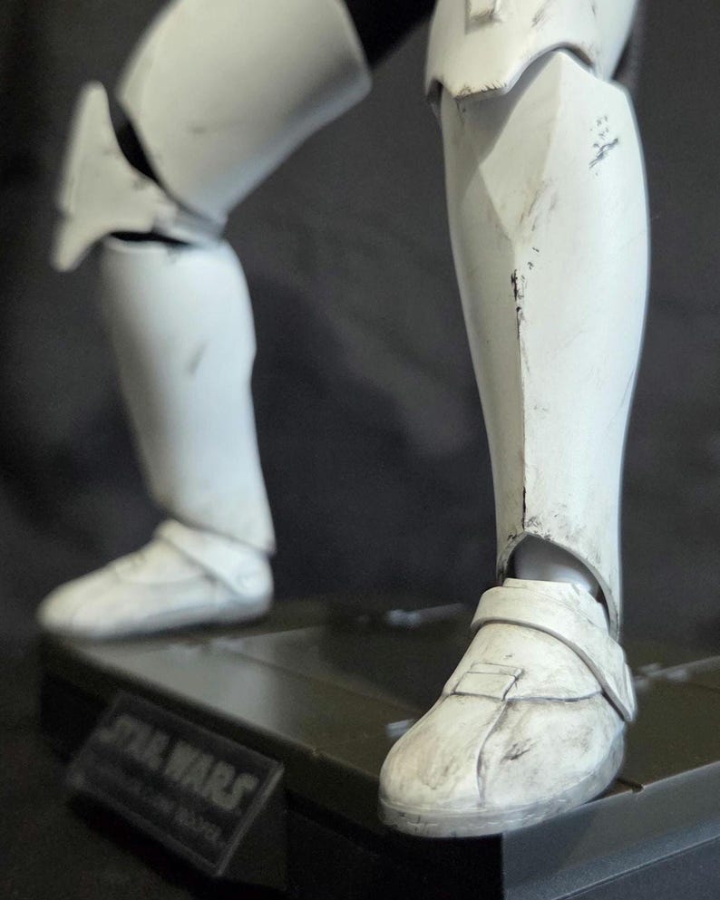 May include: Close-up of a white Stormtrooper action figure's legs and feet. The figure is detailed with battle-worn markings and is posed on a black base. The base has the Star Wars logo.