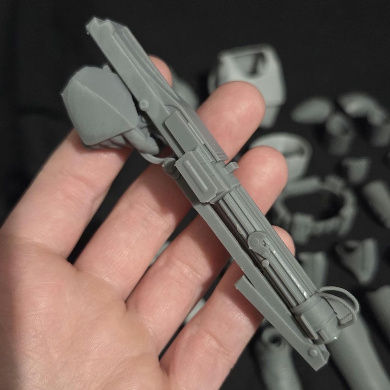 May include: A gray, plastic, mechanical component, possibly a model or part of a larger assembly. The piece has a complex design with multiple sections, including a cylindrical element and a rectangular structure. Other gray components are visible in the background.