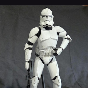 May include: A white and black action figure of a Star Wars Clone Trooper, standing on a green base. The figure is holding a black blaster rifle. The trooper is wearing white armor with black accents and a helmet.