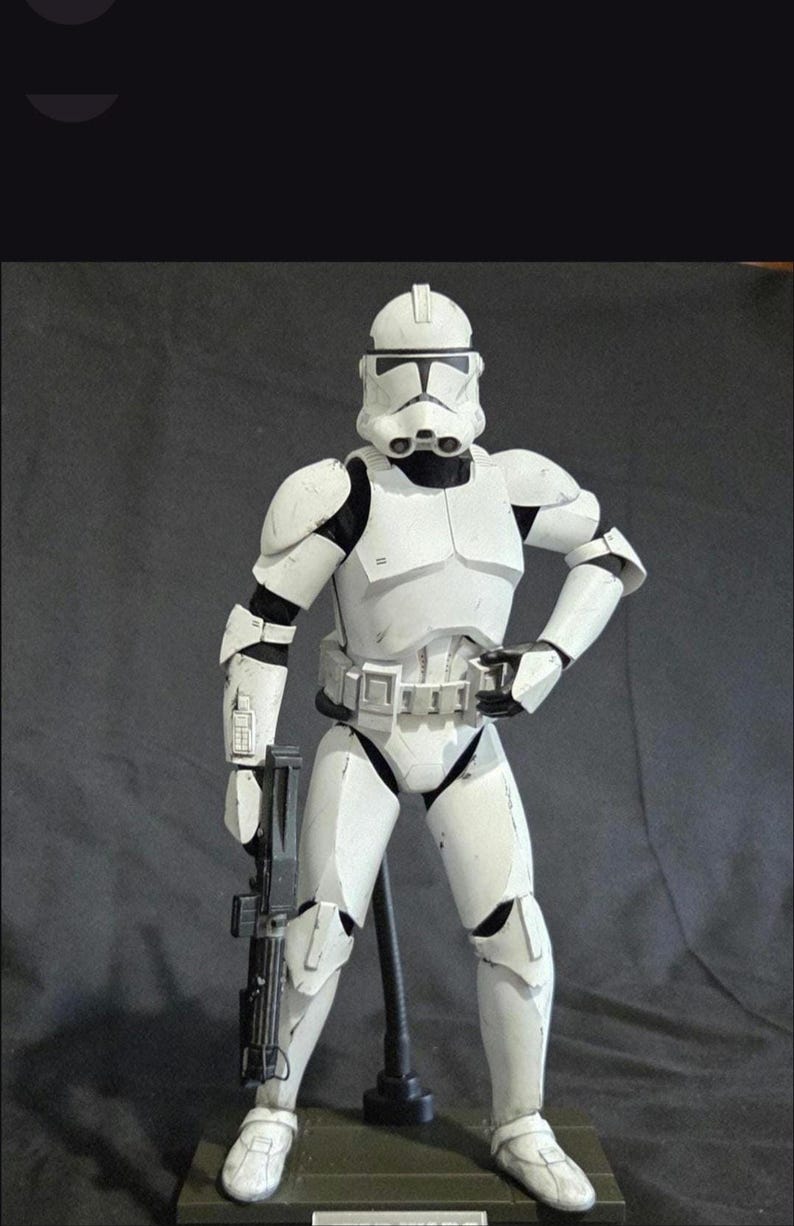 May include: A white and black action figure of a Star Wars Clone Trooper, standing on a green base. The figure is holding a black blaster rifle. The trooper is wearing white armor with black accents and a helmet.