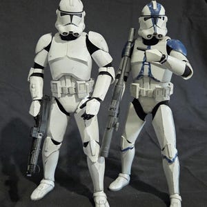 May include: Two white and black action figures of clone troopers, each holding a blaster rifle. One figure has blue accents on the armor. The figures are standing on a dark surface.