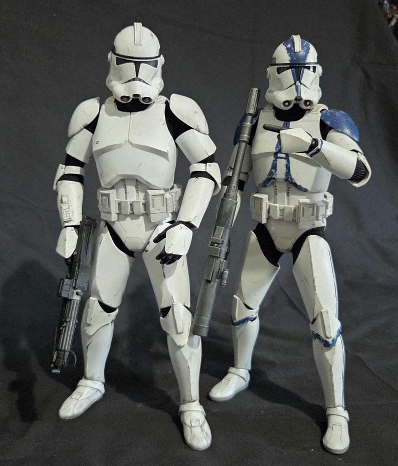 May include: Two white and black action figures of clone troopers, each holding a blaster rifle. One figure has blue accents on the armor. The figures are standing on a dark surface.