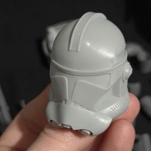 May include: A gray, plastic helmet with a distinctive design, featuring a rounded top and angular facial details. The helmet has a smooth surface and appears to be a replica or model of a character's headgear. The helmet is held in a hand.