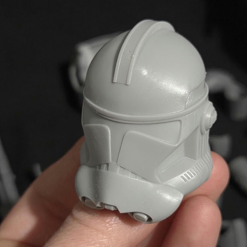 May include: A gray, plastic helmet with a distinctive design, featuring a rounded top and angular facial details. The helmet has a smooth surface and appears to be a replica or model of a character's headgear. The helmet is held in a hand.