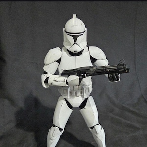 May include: A white action figure of a clone trooper from Star Wars, holding a black blaster rifle. The figure is in a combat stance, wearing white armor with black accents. The figure is approximately 12 inches tall.