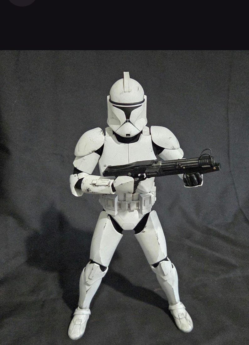 May include: A white action figure of a clone trooper from Star Wars, holding a black blaster rifle. The figure is in a combat stance, wearing white armor with black accents. The figure is approximately 12 inches tall.