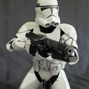May include: A white and black action figure of a clone trooper from a science fiction film. The figure is holding a black blaster rifle. The trooper is wearing white armor with black accents and a helmet. The figure is standing on a black stand.