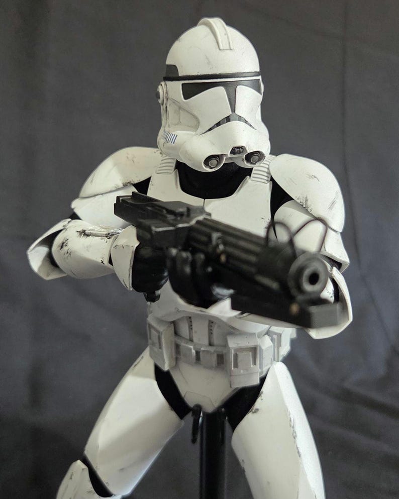 May include: A white and black action figure of a clone trooper from a science fiction film. The figure is holding a black blaster rifle. The trooper is wearing white armor with black accents and a helmet. The figure is standing on a black stand.
